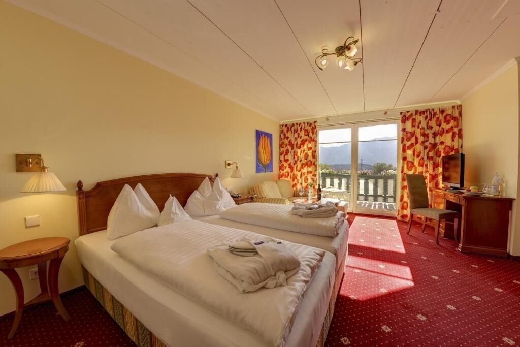 A hotel room with two beds, a desk, a chair, a TV, and a balcony with a view.