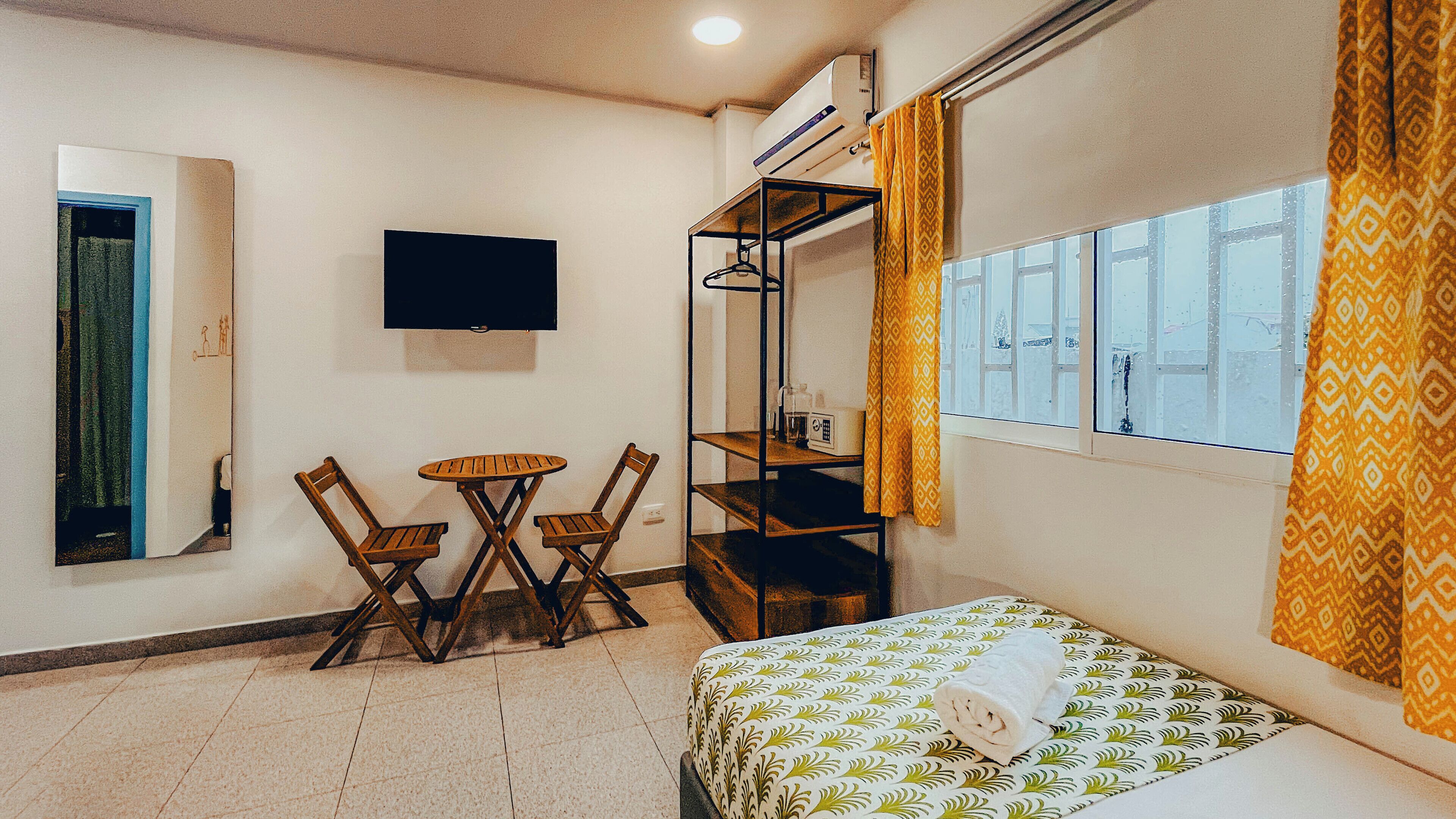A compact room with a bed, a small table with chairs, a TV mounted on the wall, and a window with curtains.