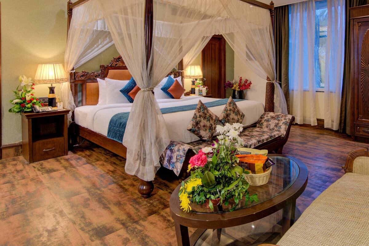 A four-poster canopy bed with a wooden frame and sheer curtains, a bedside table with a lamp, and a floral arrangement on a glass table.