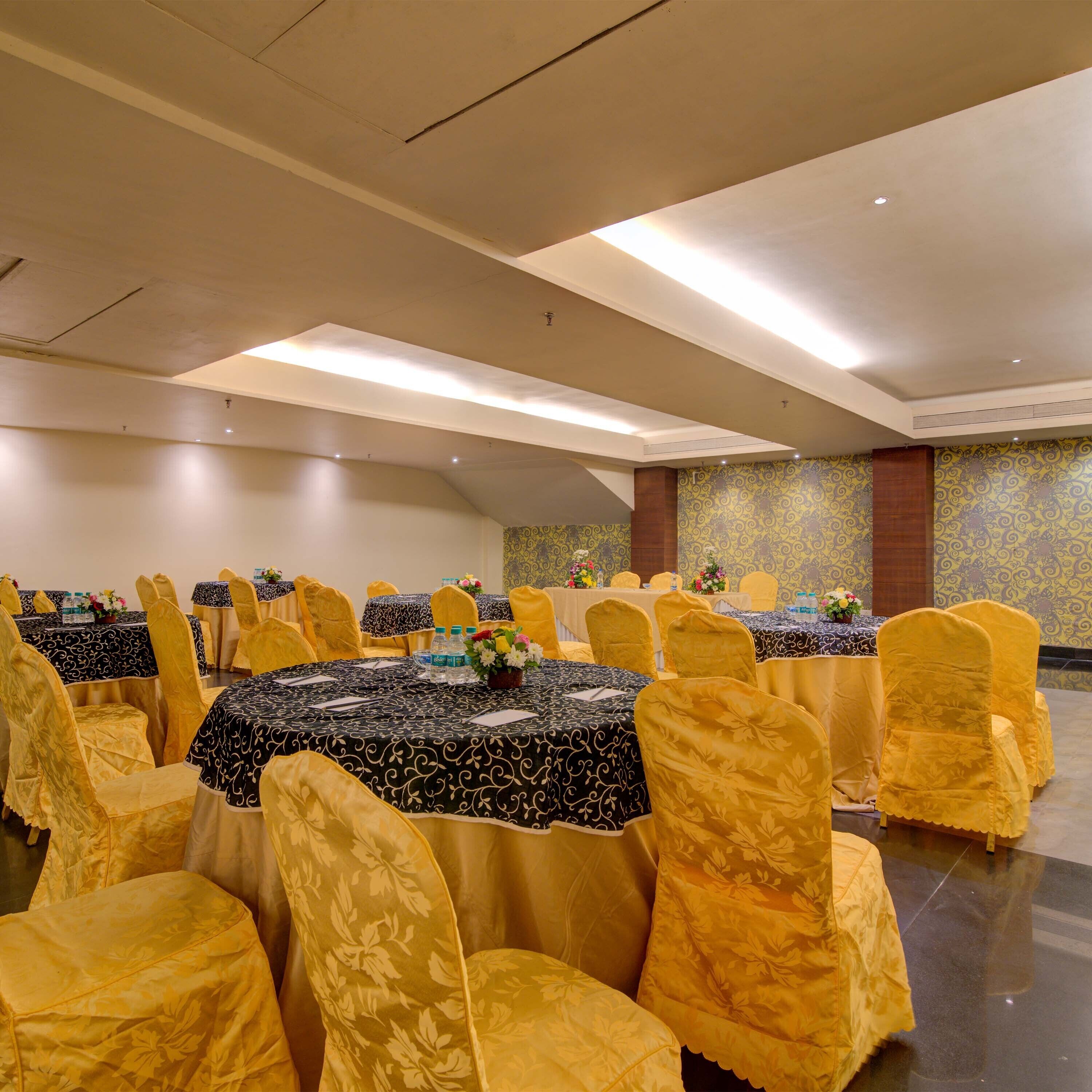 A banquet hall with round tables covered in black and white patterned tablecloths, yellow chairs with floral designs, and yellow walls with a decorative pattern.