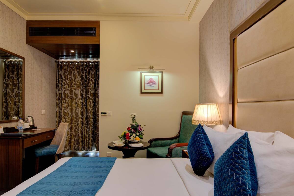 A hotel room with a bed, a desk, a chair, a table with a vase of flowers, and a wall-mounted picture.