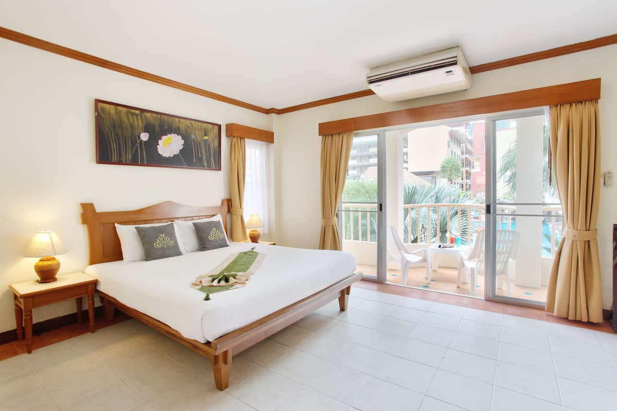 A hotel room with a large bed, a painting on the wall, a balcony with chairs, and an air conditioning unit.