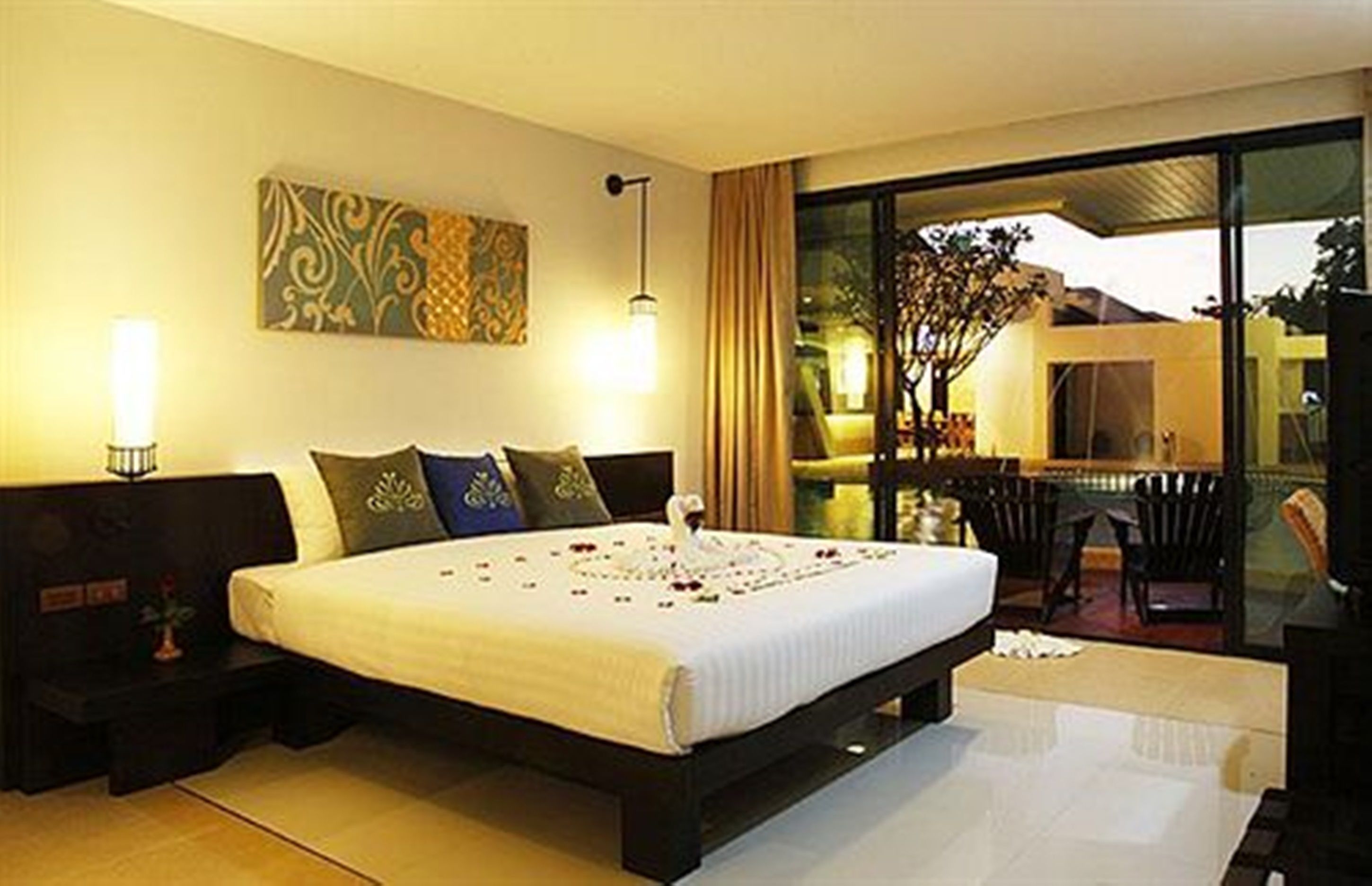 A modern bedroom with a large bed, a nightstand, a wall-mounted lamp, and a view of the outdoors through a glass door.