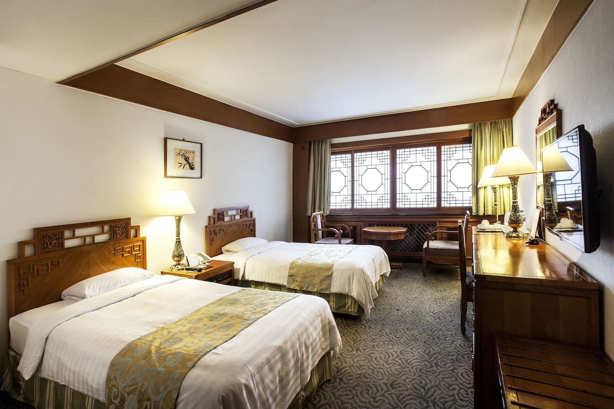 A hotel room with two beds, a desk, a chair, a lamp, and a window with decorative glass.