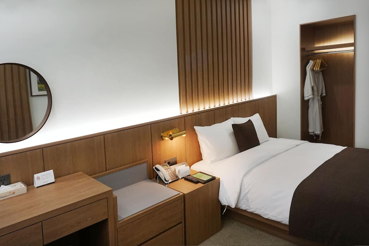 A hotel room with a wooden headboard, a bedside table with a telephone and a notepad, a bedside lamp, and a wardrobe with a robe hanging.