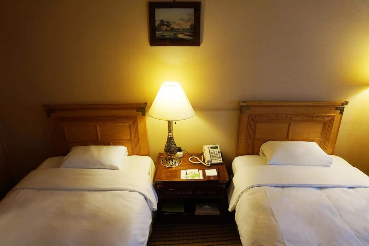 A hotel room with two single beds, a bedside table with a lamp, a phone, and a painting on the wall.