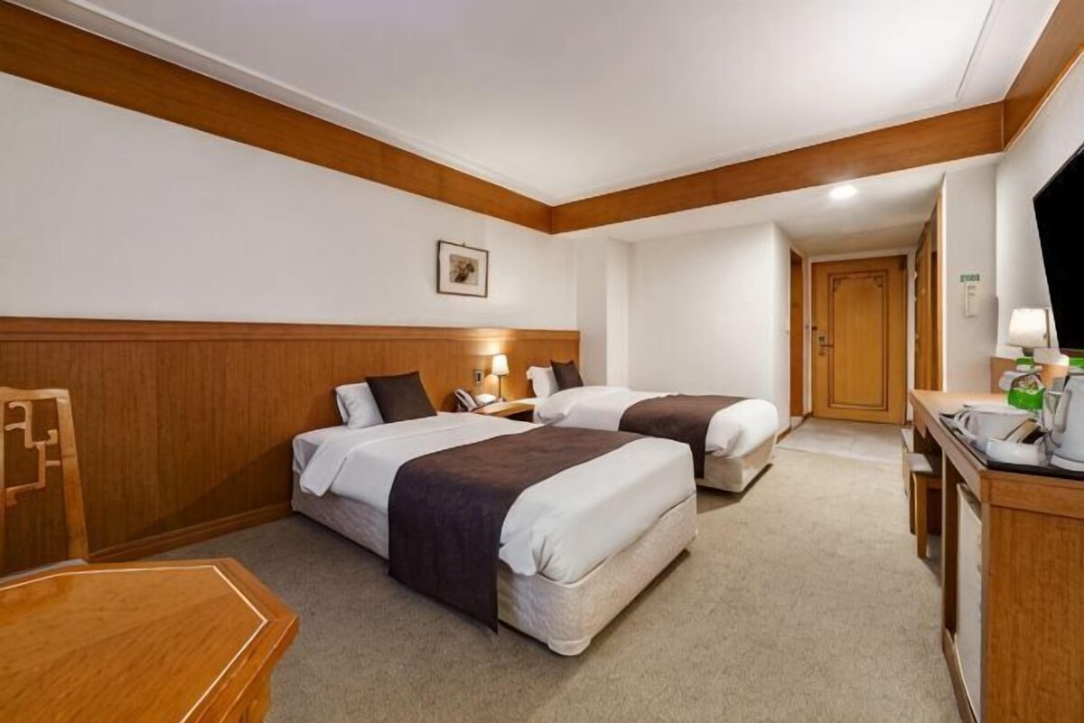 A hotel room with two beds, a wooden headboard, a desk, a chair, and a television.