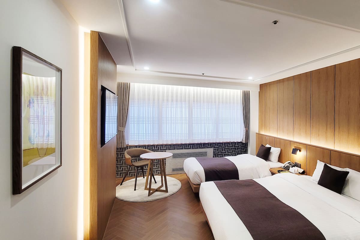 A hotel room with two beds, a desk, a chair, a window with blinds, and a framed picture on the wall.