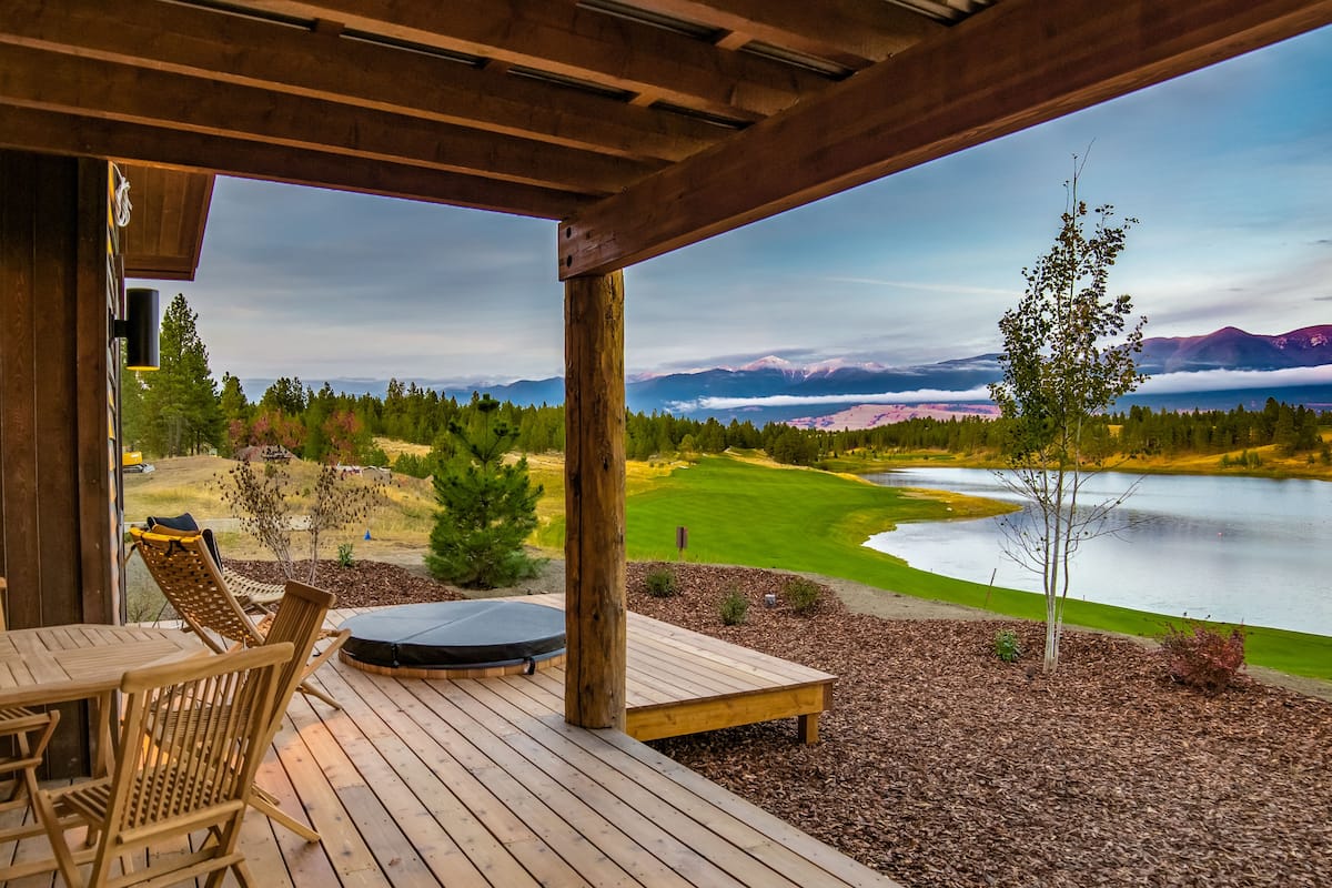 A wooden deck with a table and chairs, overlooking a lake and a golf course with mountains in the background.
