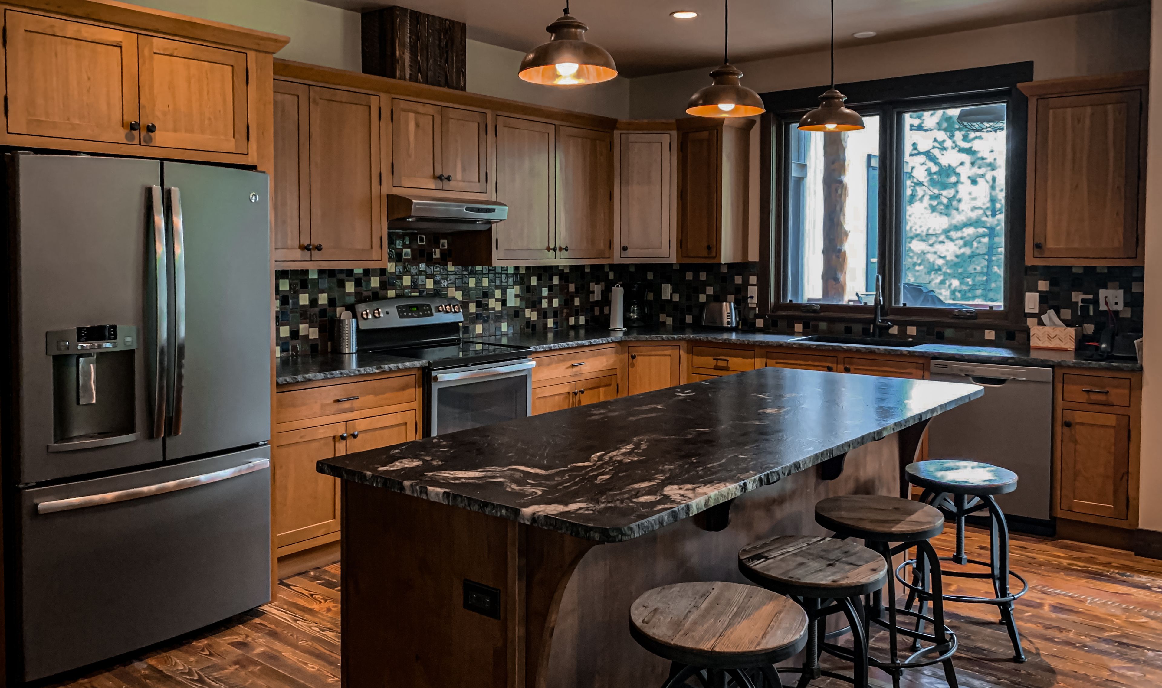 A modern kitchen with wooden cabinets, a stainless steel refrigerator, a built-in oven, and a large marble island with bar stools.