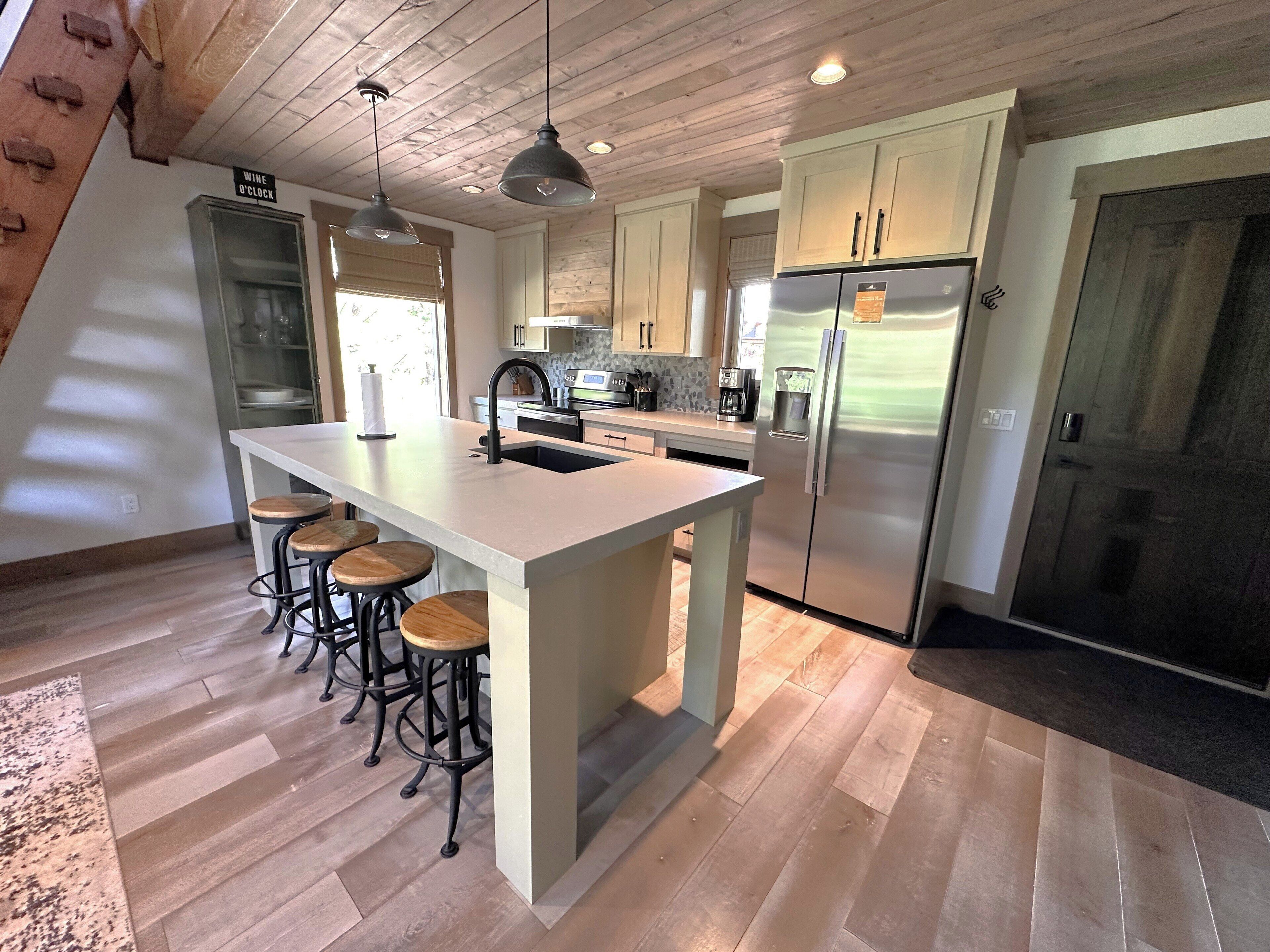Una cocina moderna con una isla central, taburetes de bar, electrodomésticos de acero inoxidable y suelo de madera.