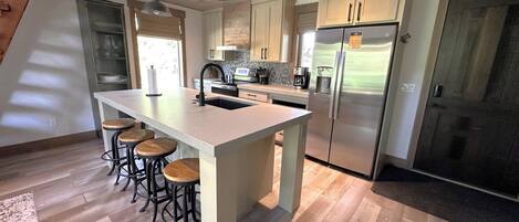 Una cocina moderna con una isla central, taburetes de bar, electrodomésticos de acero inoxidable y suelo de madera.