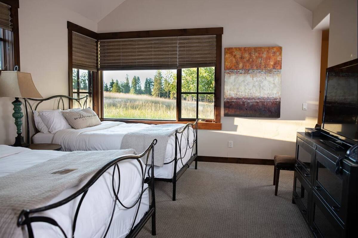 A bedroom with two beds, a large window with a view of trees, a painting on the wall, and a TV on a dresser.
