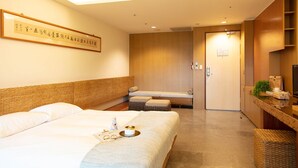 A hotel room with a large bed, a smaller bed, a sofa, a desk with a computer, and a bathroom visible through an open door.