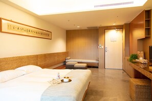 A hotel room with a large bed, a smaller bed, a sofa, a desk with a computer, and a bathroom visible through an open door.