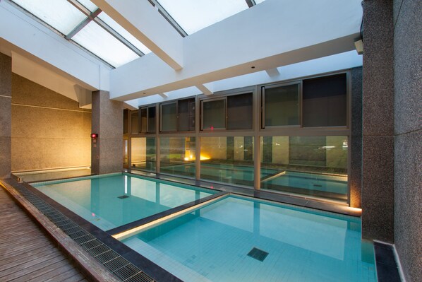 An indoor swimming pool with clear glass walls, a tiled floor, and a wooden deck area.