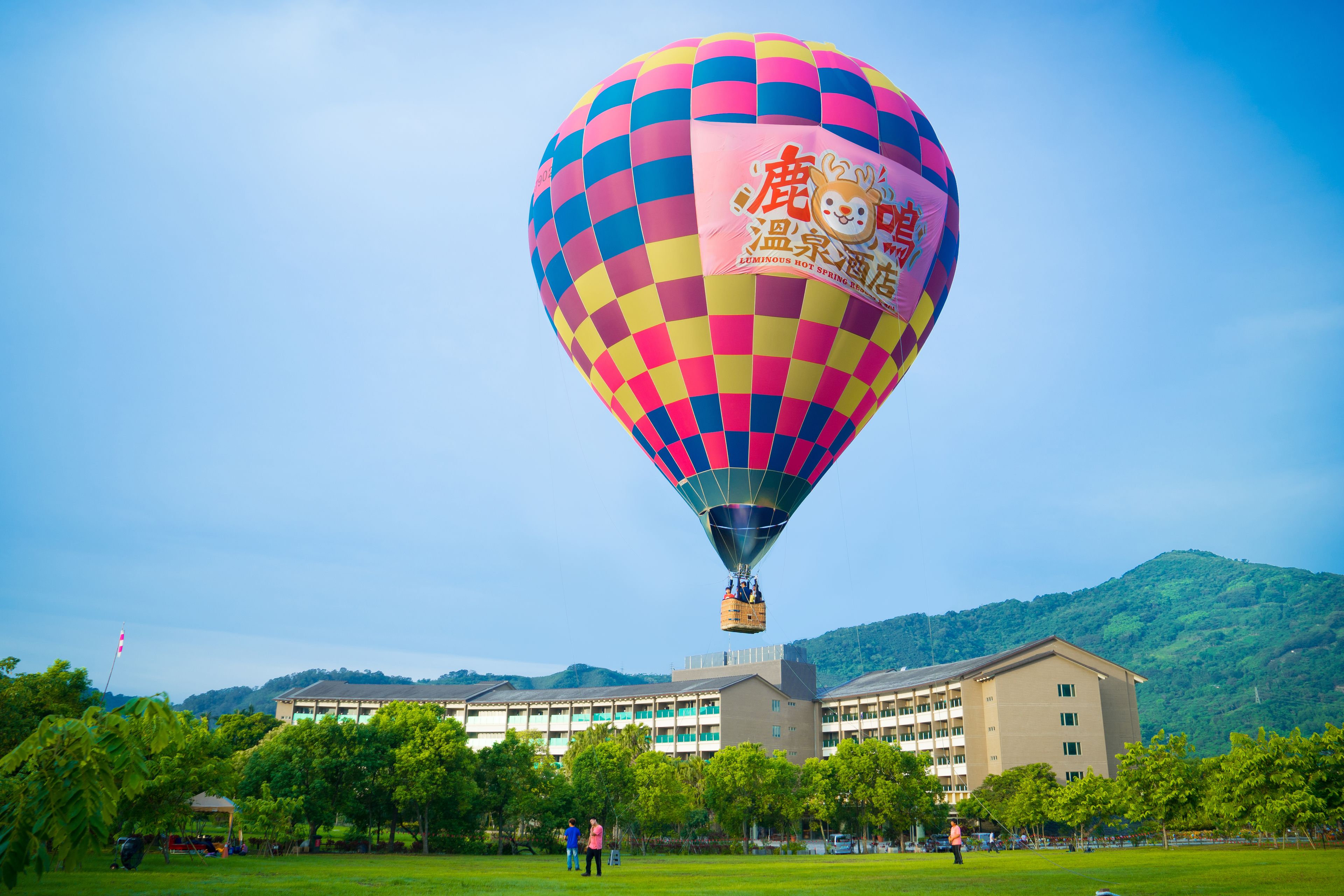 A hot air balloon with a checkered pattern and a logo, flying over a building surrounded by trees.