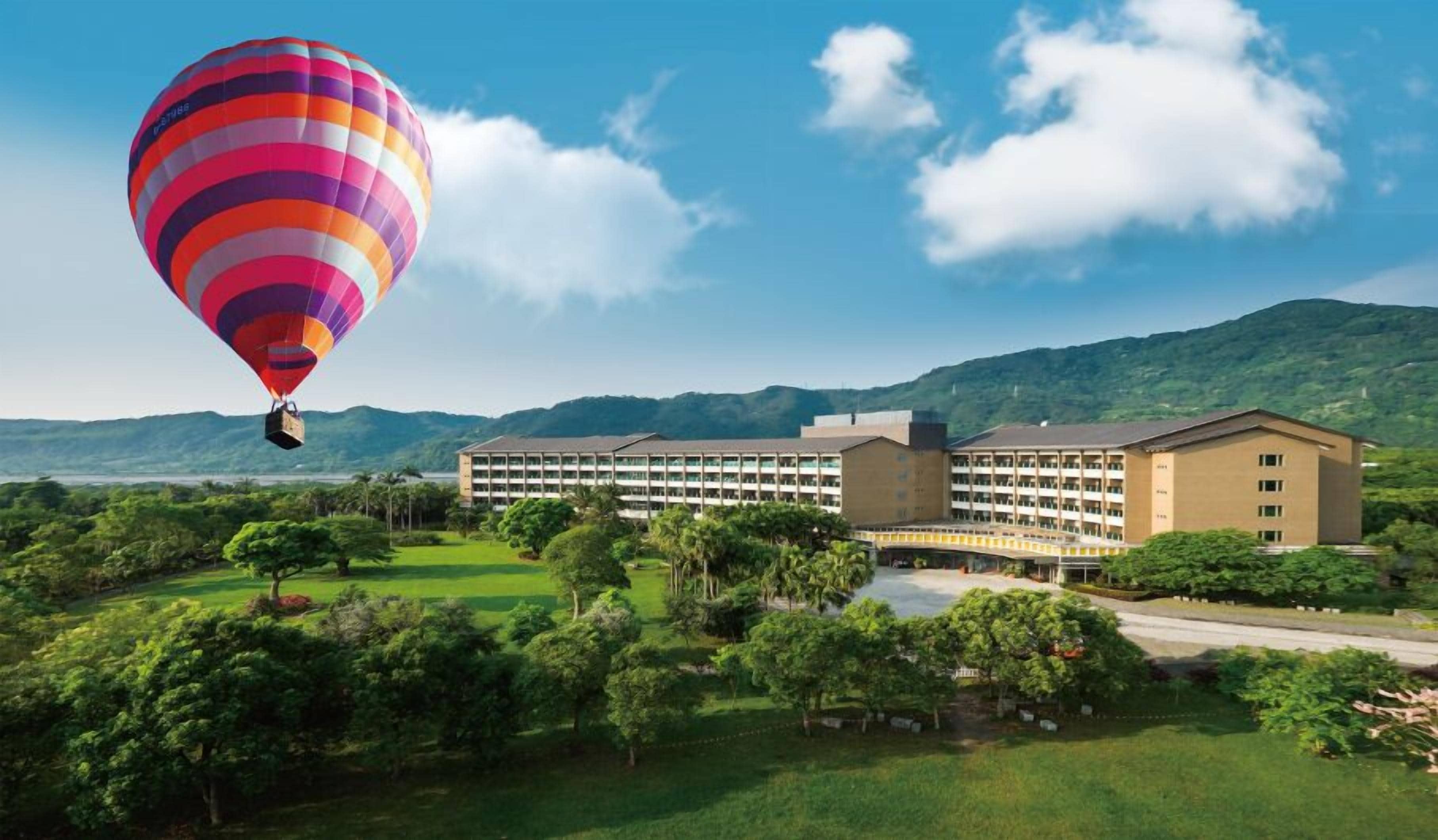 A large hotel with multiple floors and a central courtyard, surrounded by lush greenery and mountains.
