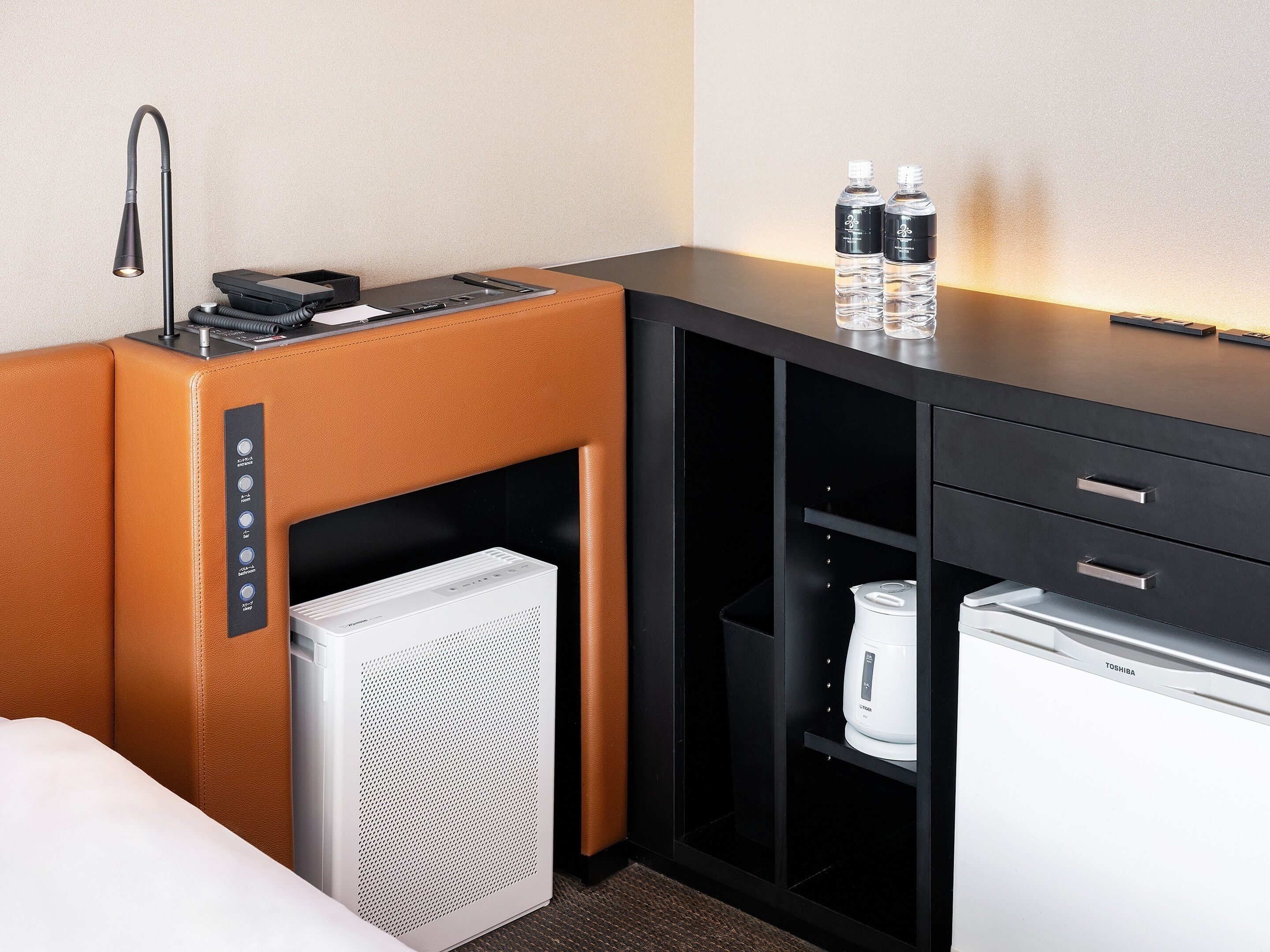 Habitación de hotel con cama, minibar, refrigerador, cafetera y escritorio con lámpara y botellas de agua.