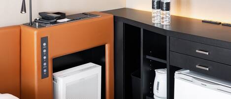 Habitación de hotel con cama, minibar, refrigerador, cafetera y escritorio con lámpara y botellas de agua.