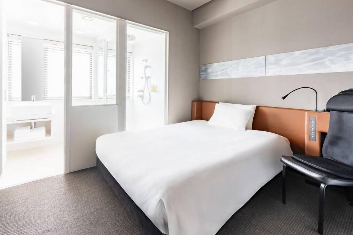 Habitación de hotel con cama, una silla, baño con ducha y vista al exterior.
