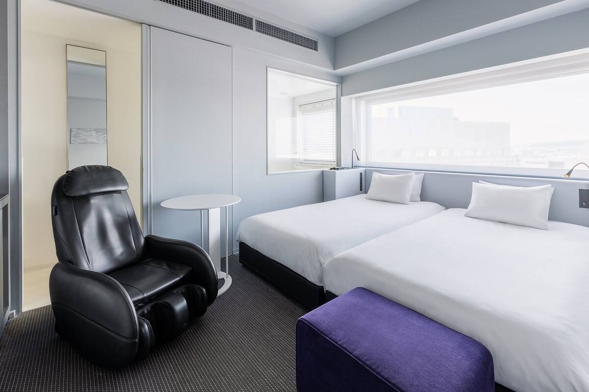 A modern hotel room with a bed, a massage chair, a small round table, and a window with a city view.