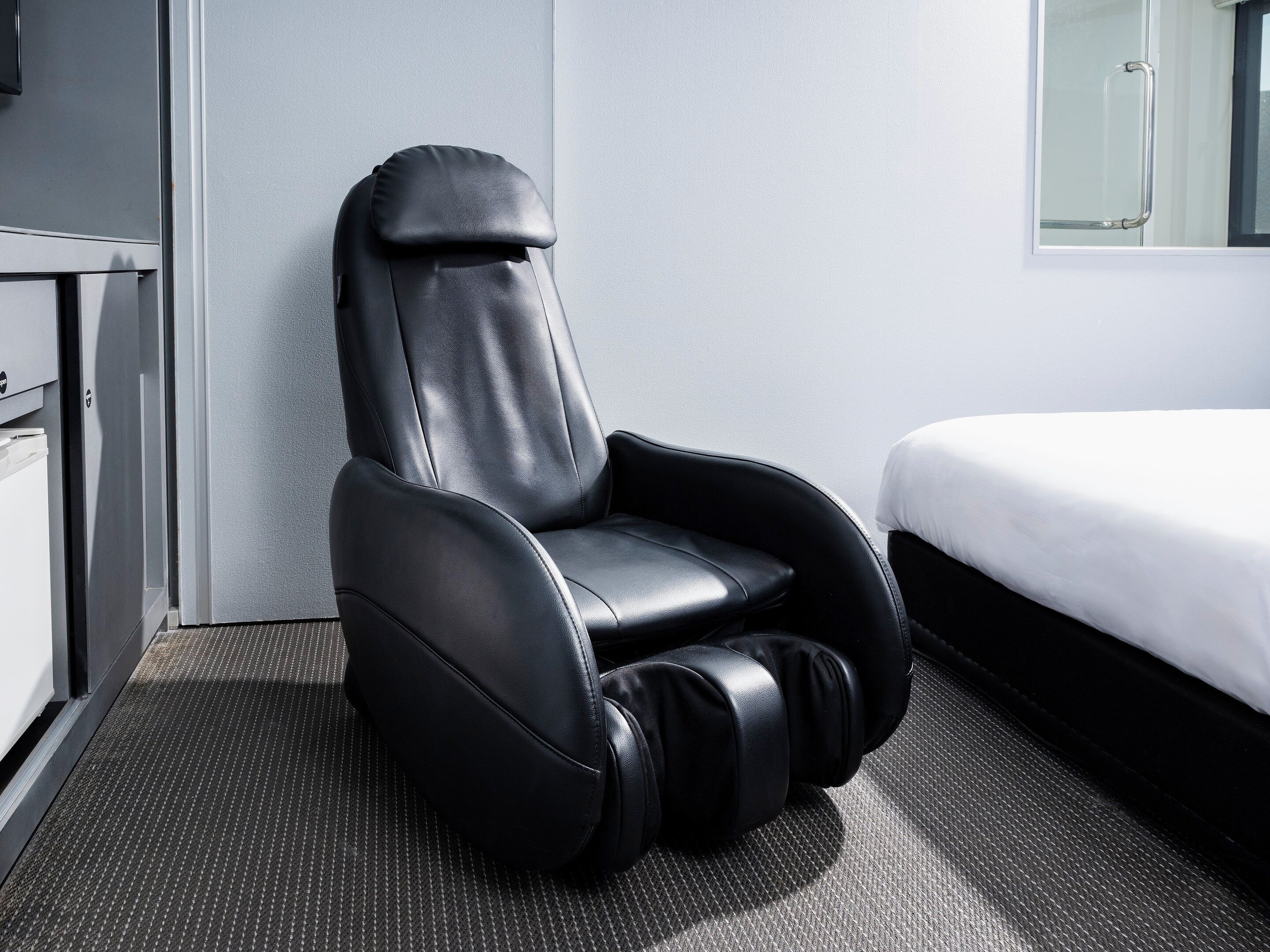 A black massage chair in a hotel room with a bed and a small desk.