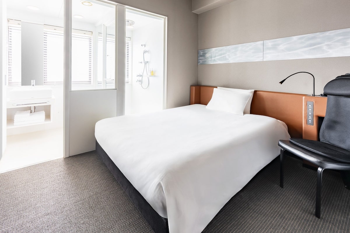 A hotel room with a bed, a chair, a shower area, and a bathroom.