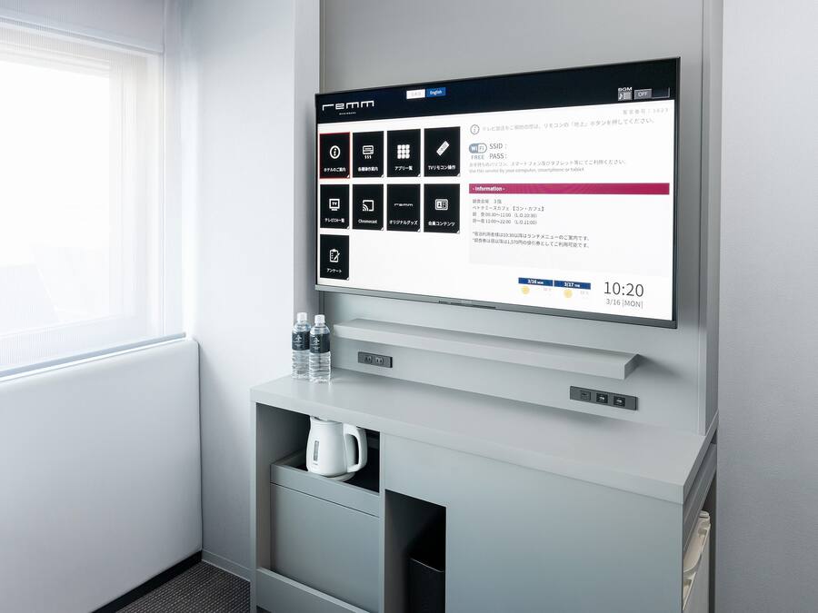 Hotellihuone, jossa on seinälle asennettu televisio, joka näyttää tietoja huoneesta, vesipullo, vedenkeitin ja latausasema.