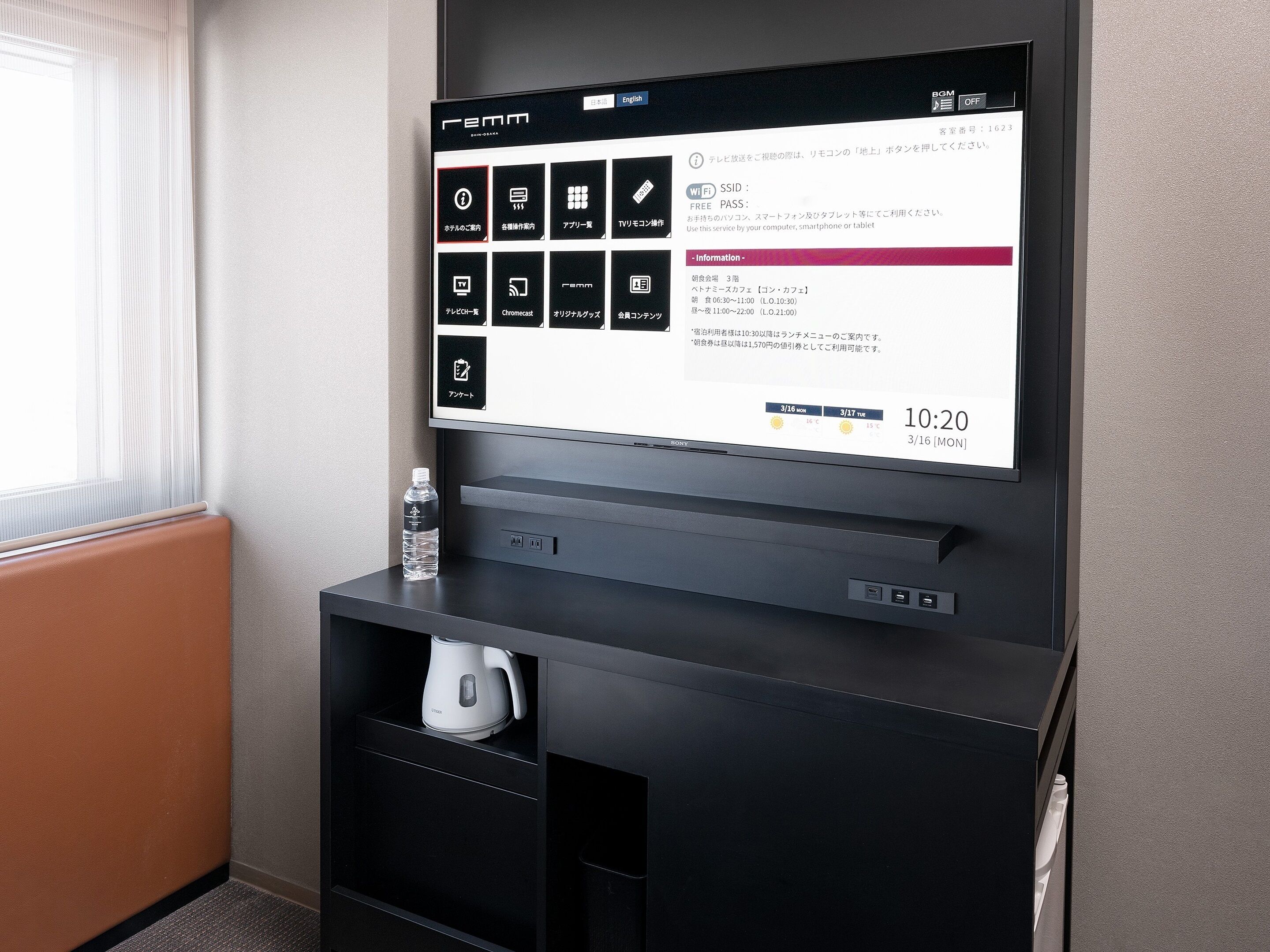 Ett hotellrum med en platt-TV som visar hotellets informationssystem, en vattenflaska och en vit vattenkokare placerad på ett svart skåp.