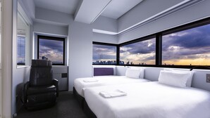 Ett modernt hotellrum med en stor säng, en massagefåtölj och utsikt över staden genom stora fönster.