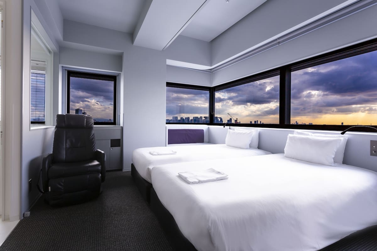 A modern hotel room with a large bed, a massage chair, and a city view through large windows.