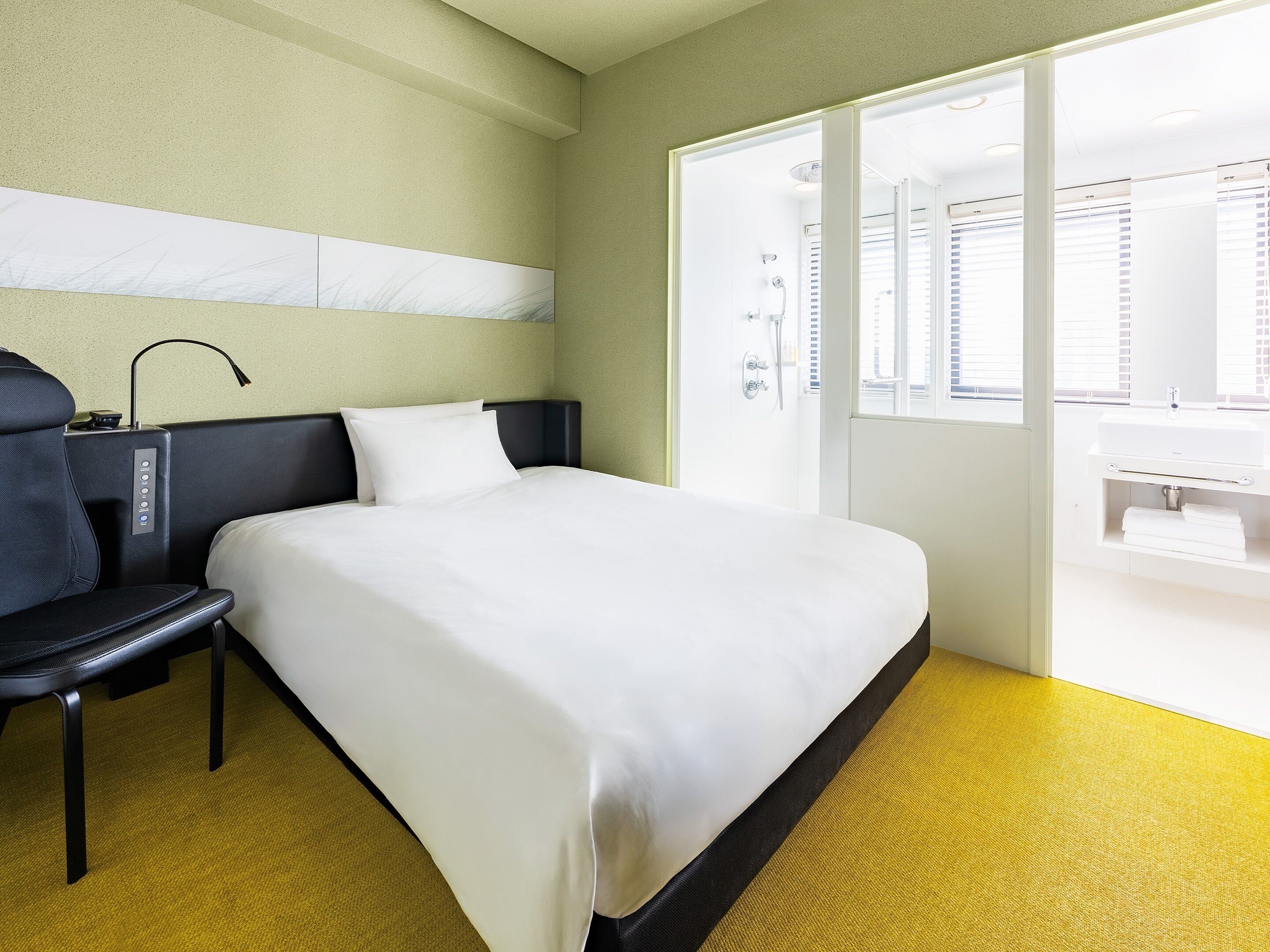 A hotel room with a bed, a desk, a chair, a window, and a bathroom visible through the door.