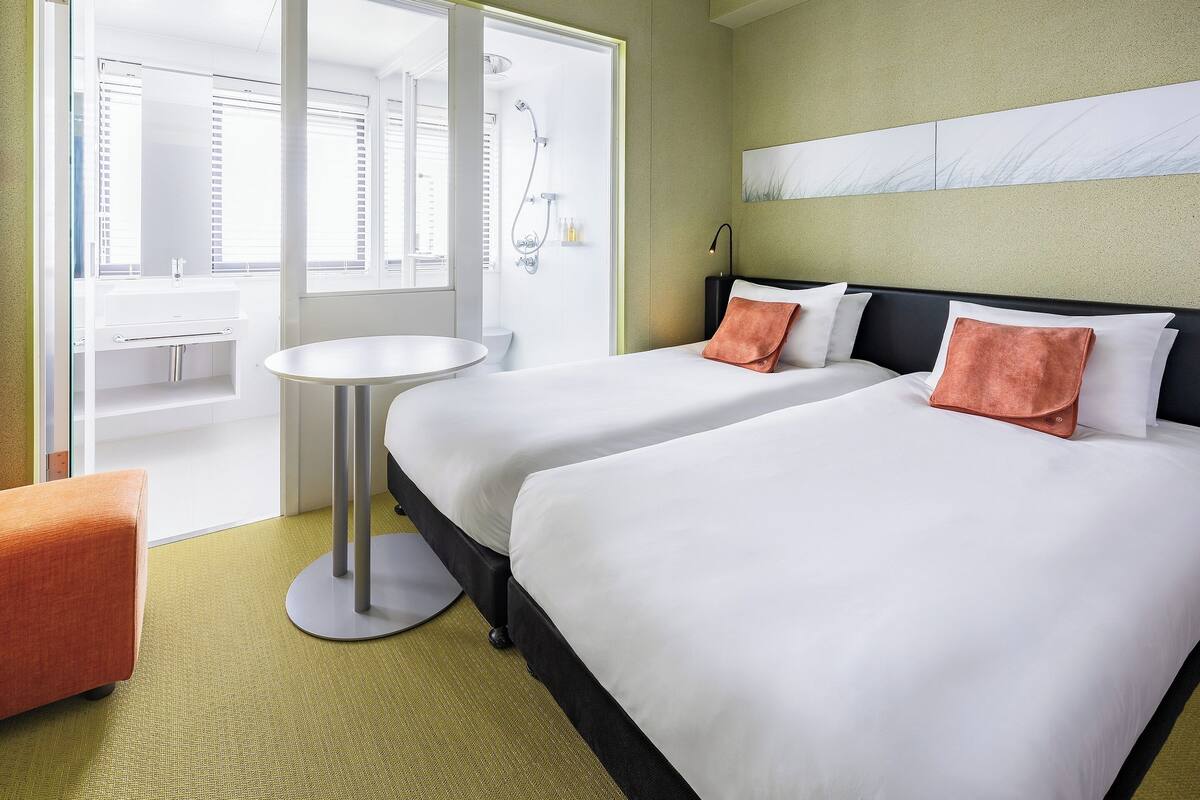A hotel room with two beds, a round table, a bathroom with a sink and shower, and a view of the city.
