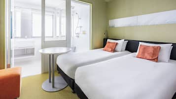 A hotel room with two beds, a round table, a bathroom with a sink and shower, and a view of the city.