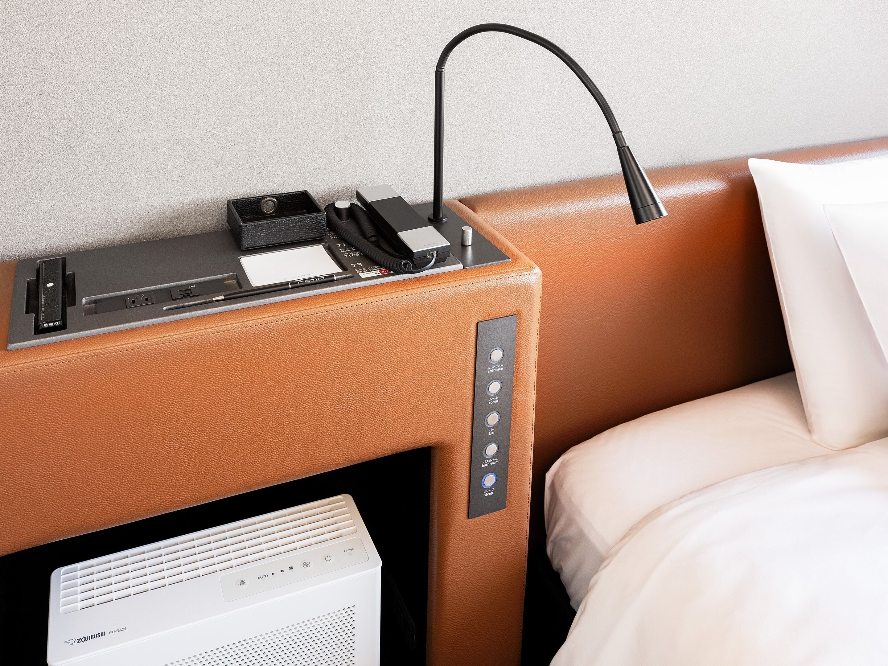 A hotel room with a bed, a control panel with buttons, and a white device on the floor.