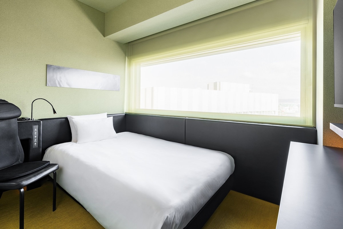A hotel room with a bed, a desk, a chair, a TV, and a window with blinds.
