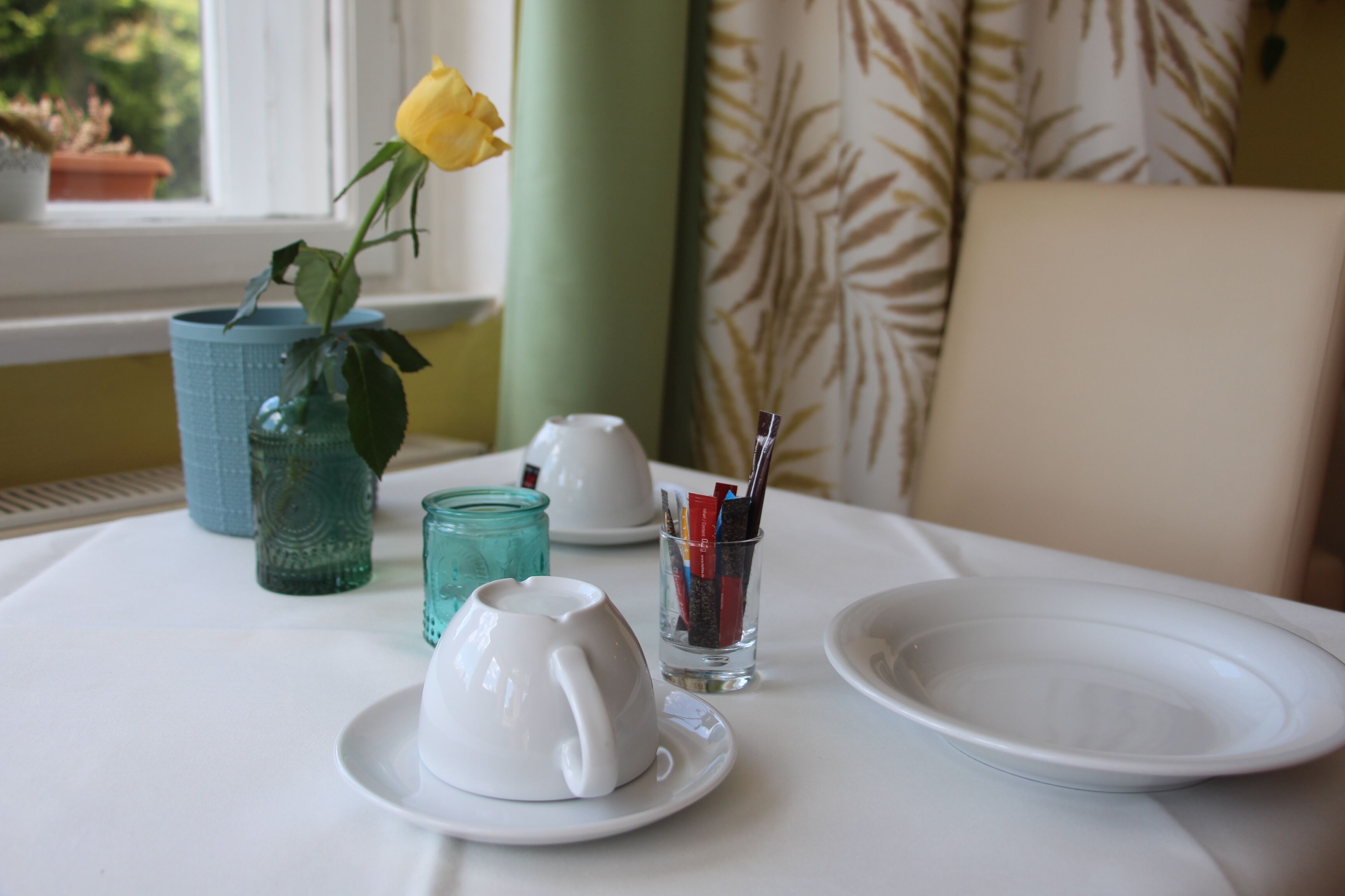 Une table à manger avec une assiette blanche, une tasse blanche, un verre contenant des couverts et un vase bleu avec une fleur jaune.