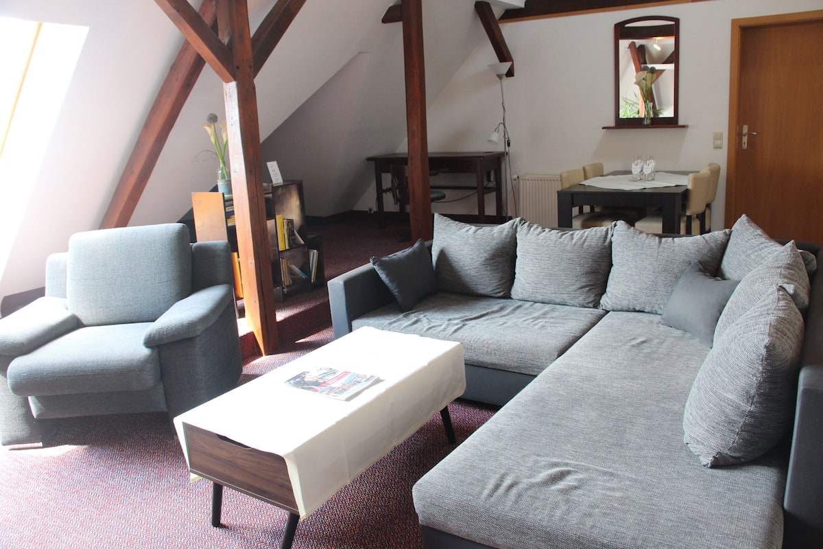 Un salon avec un canapé gris, un fauteuil assorti et une petite table. On aperçoit une table à manger avec des chaises au fond de la pièce.