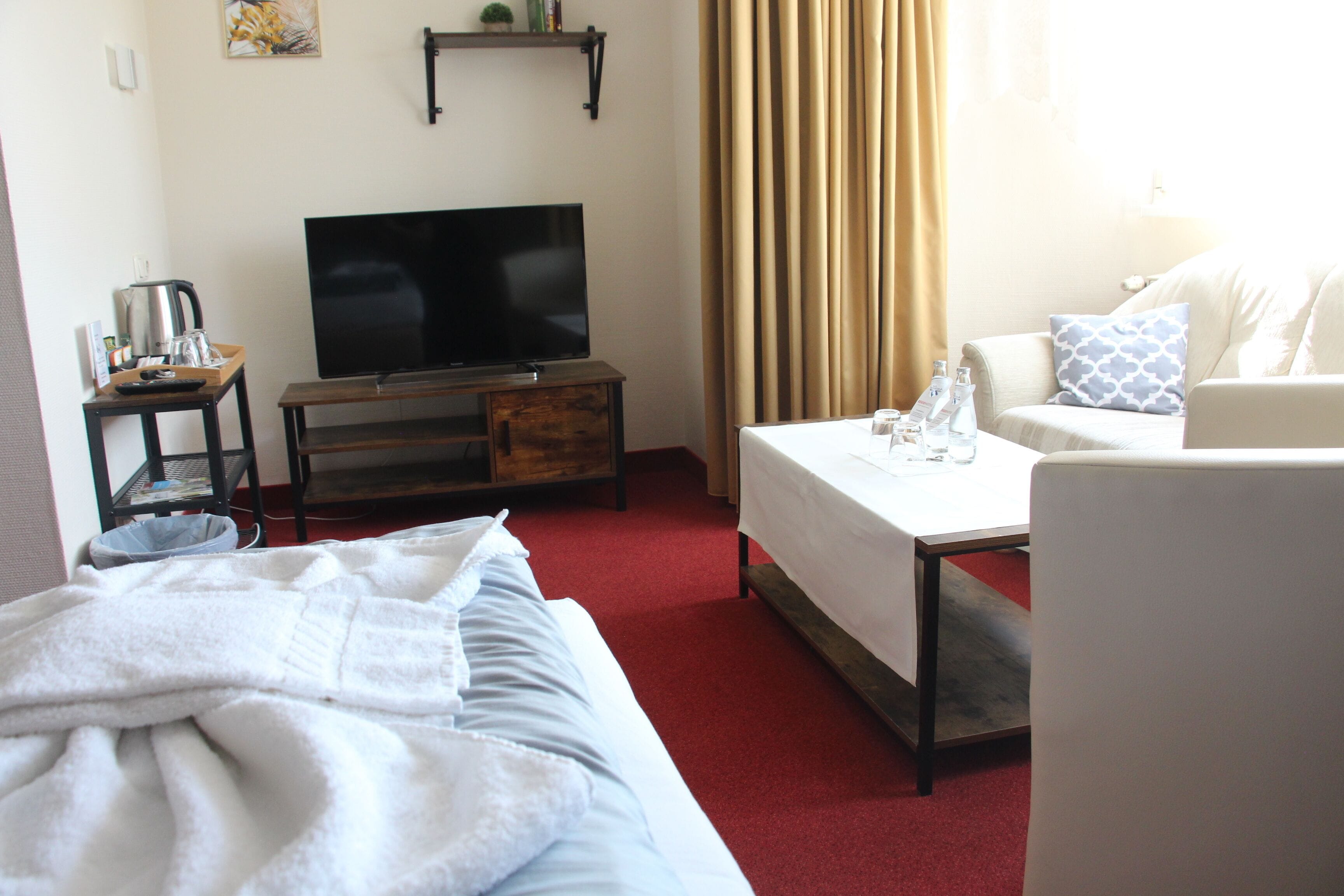 Habitación de hotel con cama, televisor sobre un soporte de madera, mesa de centro, sofá y ventana con cortinas.