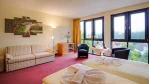 Habitación de hotel con cama, sofá, sillón, mesita con un florero y ventana con vistas.