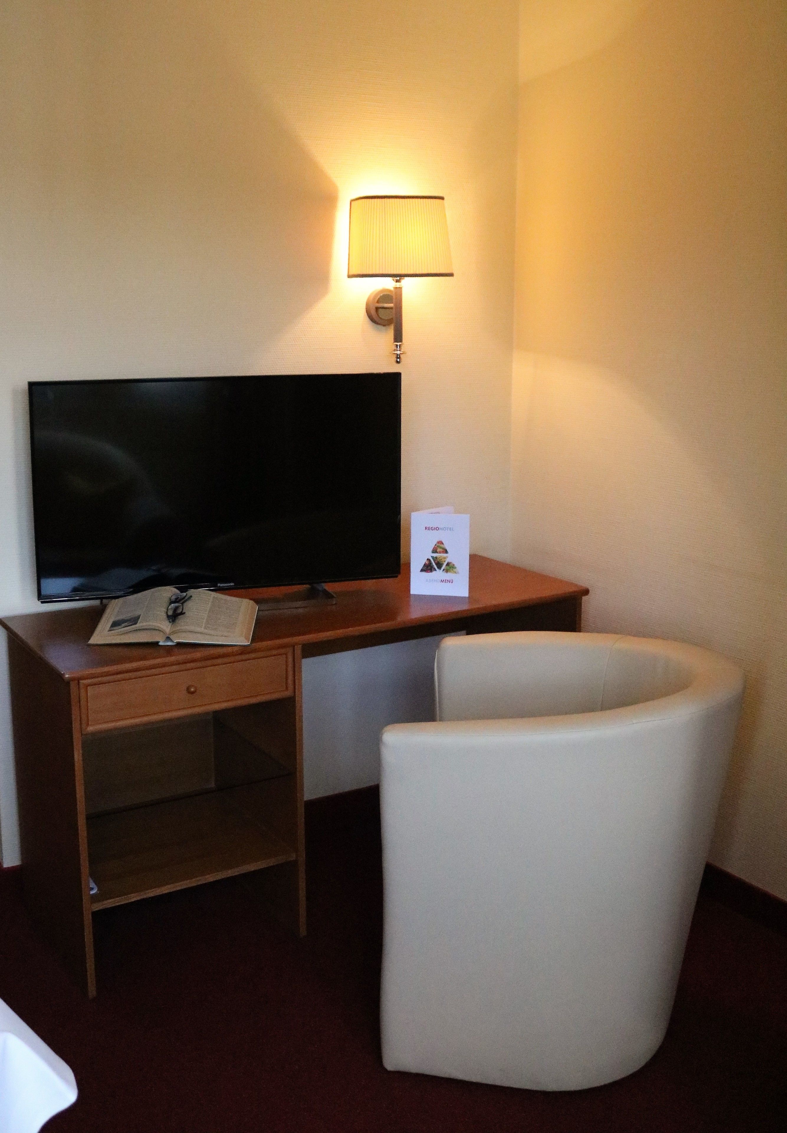 A hotel room with a flat-screen TV on a wooden desk, a white armchair, a lamp, and a small card on the desk.
