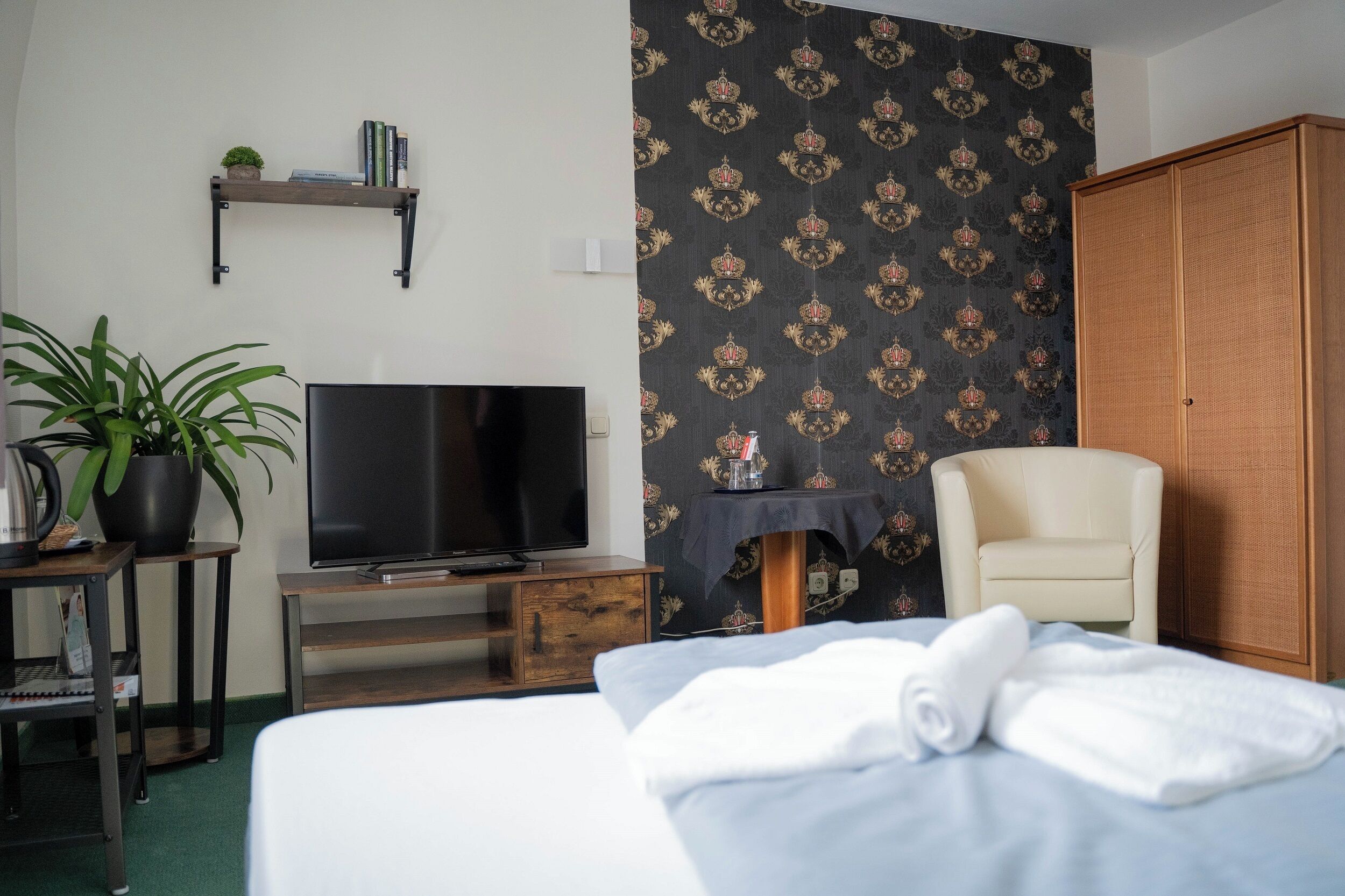 Ein Hotelzimmer mit Bett, Fernseher, Sessel, einem kleinen Tisch mit Pflanze und einem Kleiderschrank.