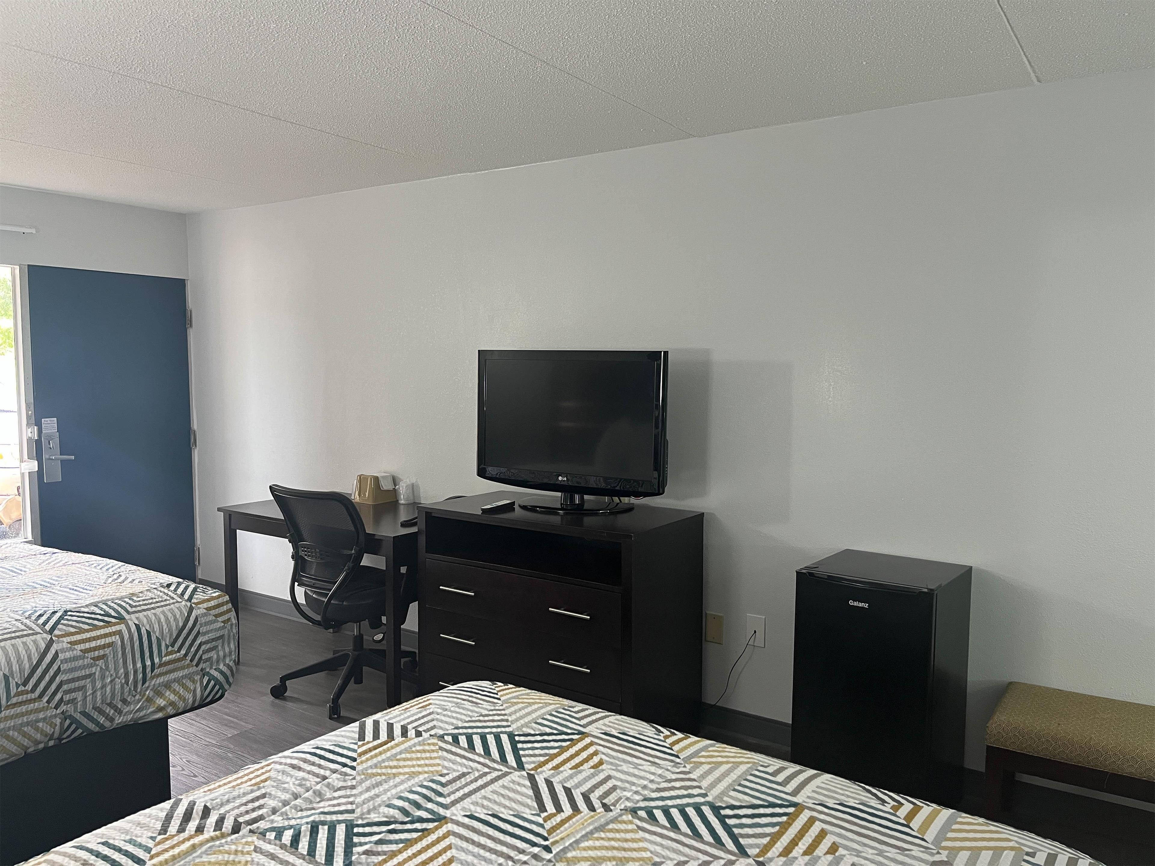 A hotel room with two beds, a desk with a computer and TV, a chair, and a small bench.