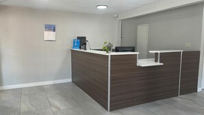 A reception desk with a dark wood finish, a white countertop, and a small plant. There is a sign on the wall and a security camera above the desk. - Motel 6 – Spindale, NC – Forest City (Spindale)