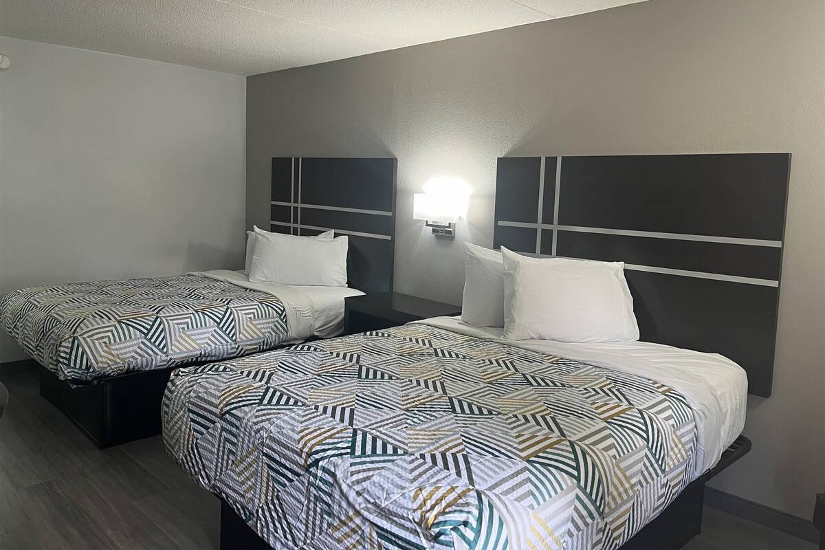 A hotel room with two beds, each with a patterned bedspread and white pillows. There is a wall-mounted light fixture above the beds.