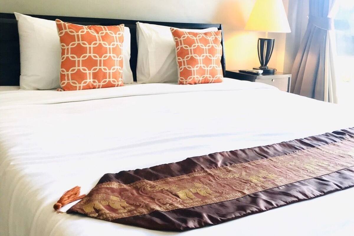 Ein Hotelzimmer mit einem großen Bett, zwei gemusterten Kissen, einem gerahmten Bild an der Wand, einer Nachttischlampe und einem Fenster mit Vorhängen.