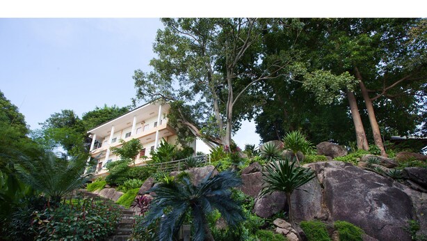 Un edificio de varios pisos con balcón, rodeado de abundante vegetación y grandes rocas.