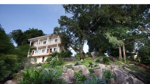 Un edificio de varios pisos con balcones, rodeado de abundante vegetación y grandes rocas.