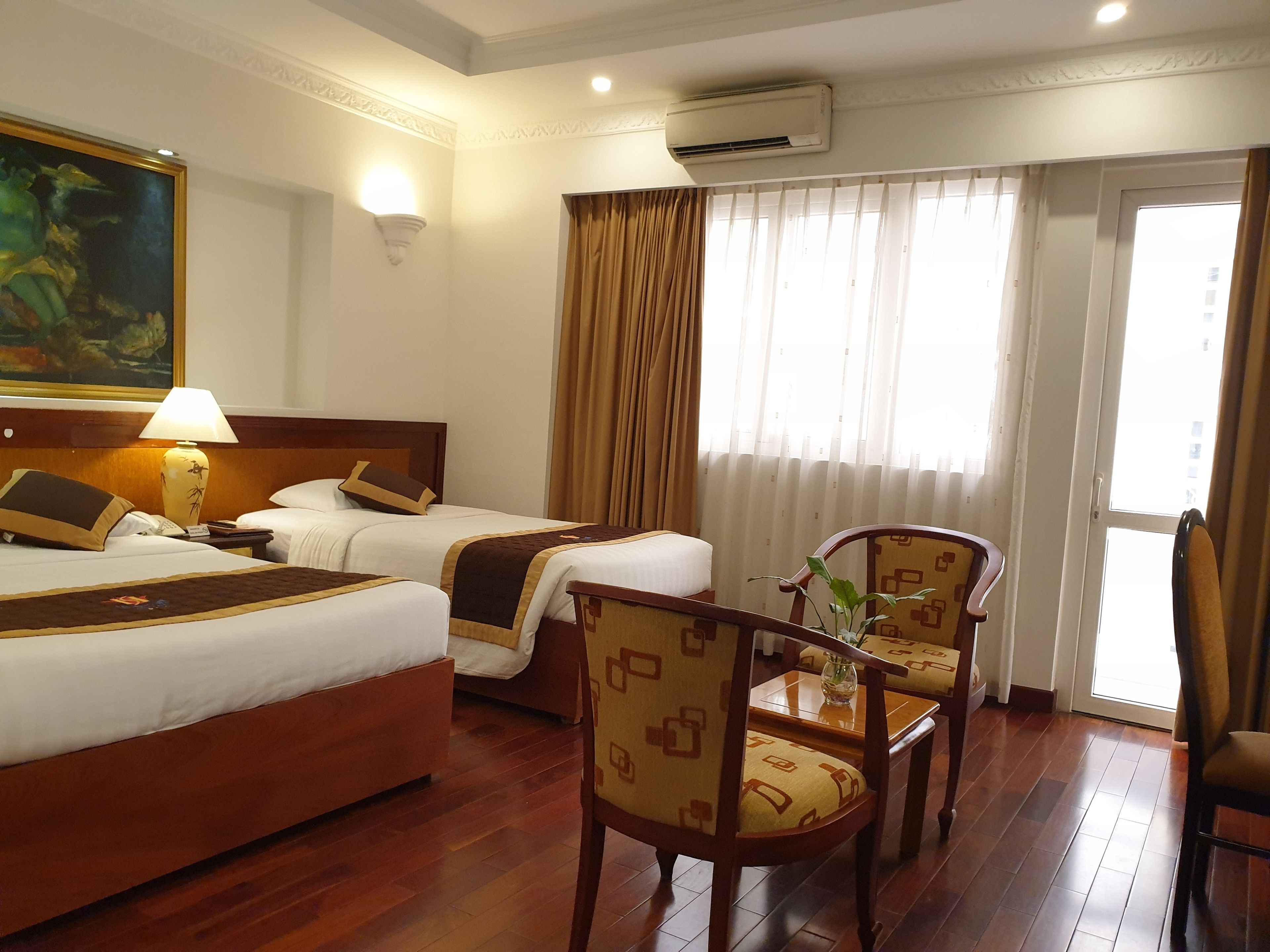 A hotel room with two beds, a wooden floor, a painting on the wall, a lamp, an air conditioner, and a window with curtains.